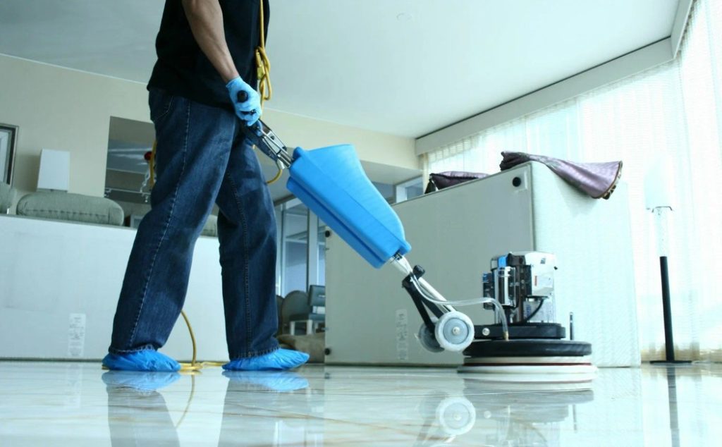 A person wearing blue gloves and shoe covers is using a floor polishing machine to clean a glossy floor, showcasing professional Floor Care Services.