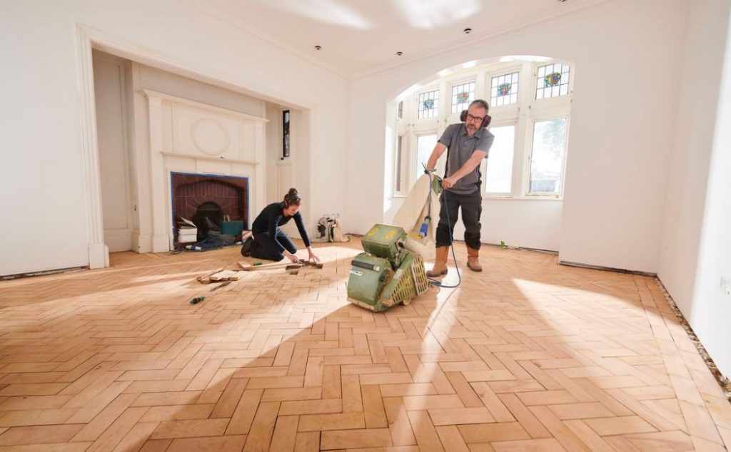 The image shows professionals working on a hardwood floor, providing floor restoration services to refinish and restore the wood's original beauty.