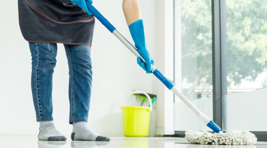 A person wearing gloves and socks is mopping a shiny floor near a large window, with a yellow bucket in the background, representing professional Floor Care Services.