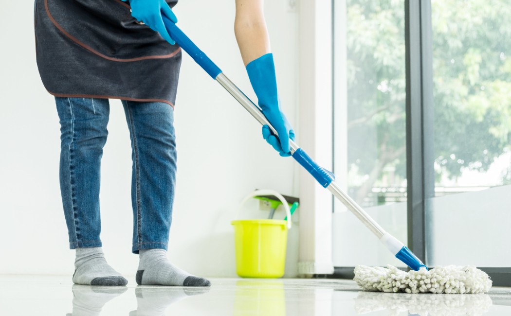 A person wearing gloves and socks is mopping a shiny floor near a large window, with a yellow bucket in the background, representing professional Floor Care Services.