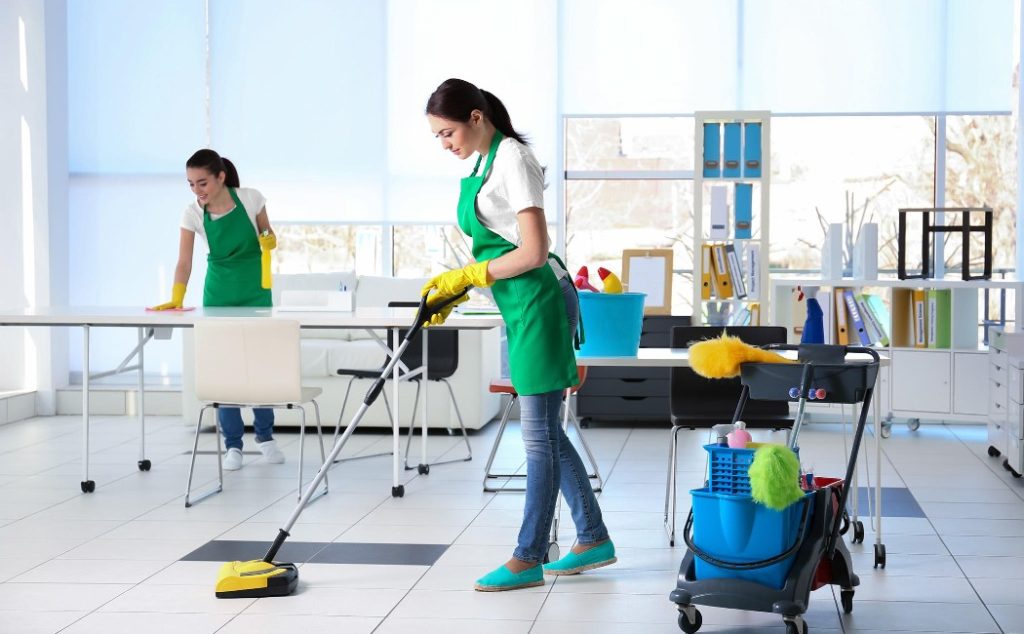 Two professionals are performing commercial cleaning services in a bright office, using cleaning equipment to maintain a spotless workspace.